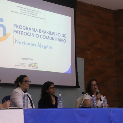 Painel com cinco pessoas sentadas à mesa com microfones e garrafas de água, diante de uma tela que exibe slide sobre o Programa Brasileiro de Patrocínio Comunitário para Nacionais Afegãos, apresentado pelo Ministério da Justiça e Segurança Pública.