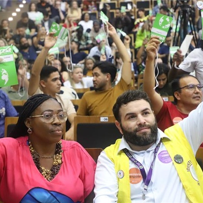 Auditório cheio de pessoas participando da COMIGRAR, todas sentadas em fileiras e levantando cartões de votação. No primeiro plano, duas pessoas seguram os cartões, uma delas usando um colete amarelo com crachá e adesivos e outra ao lado de blusa rosa também levantando um cartão.