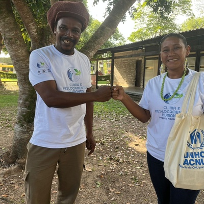 Homem negro e mulher indígena apertam as mãos, ambos olhando para a câmera. Ela carrega uma bolsa ecológica com a logomarca do ACNUR. Ao fundo existem árvores e uma casa.