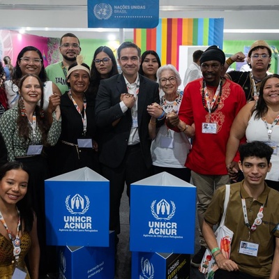Grupo de pessoas reunidas em um estande da ONU com dois totens azuis exibindo o logotipo do ACNUR (Agência da ONU para Refugiados). Ao fundo, há um painel com faixas coloridas e outro com o emblema das Nações Unidas. Algumas pessoas usam crachás e seguram sacolas, indicando participação na COP30.