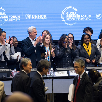 Grupo diverso de participantes aplaude no palco durante a sessão de encerramento da Revisão de Progresso do Fórum Global sobre Refugiados. Ao fundo, um painel azul exibe os logotipos do ACNUR e do Fórum Global sobre Refugiados. Em primeiro plano, representantes e delegados circulam pelo espaço da conferência, com mesas, microfones e placas institucionais visíveis, reforçando o caráter formal e internacional do evento.