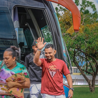 Pessoas descendo de um ônibus estacionado; uma delas acena com a mão enquanto outra caminha carregando uma criança enrolada em um cobertor. Árvores e uma área verde aparecem ao fundo.