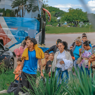 Grupo de pessoas caminhando com malas e crianças próximo a um ônibus estacionado, em uma área aberta com vegetação em primeiro plano e árvores ao fundo.