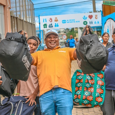 Grupo de pessoas carregando mochilas e bolsas grandes com a logomarca do ACNUR, enquanto caminham por uma área externa. Ao fundo, há um portão com a sinalização da organização ‘Aldeias Infantis SOS’. As pessoas parecem estar em movimento, algumas levantando as bagagens, com construções e um mural colorido ao redor.