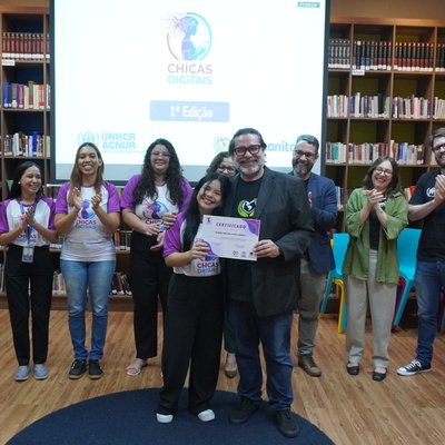 Grupo de pessoas reunidas em uma biblioteca durante um evento do programa ‘Chicas Digitais’. No centro, duas pessoas seguram um certificado. Outras pessoas ao fundo aplaudem. Atrás do grupo há uma tela de projeção exibindo o logotipo do evento e estantes cheias de livros.