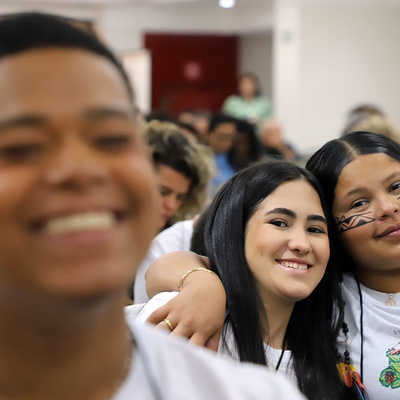 Crianças sentadas em um auditório. Em primeiro plano, uma criança aparece desfocada. Ao fundo, duas crianças próximas uma da outra olham em direção à câmera, vestindo camisetas claras com ilustrações. Há outras pessoas sentadas atrás, em ambiente interno bem iluminado.