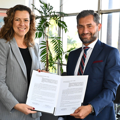 Duas pessoas em ambiente institucional seguram documento oficial aberto após assinatura, com plantas e janelas ao fundo