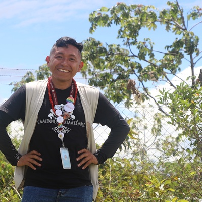 Pessoa em ambiente rural com vegetação e cerca de arame, usando colete claro, camiseta preta e crachá, em registro institucional ao ar livre.