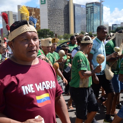Homem indígena em primeiro plano usa camiseta vinho com o texto ‘INE WARAO’ e uma pequena bandeira estampada, caminhando em meio a um grupo em marcha. Ao redor, várias pessoas vestem camisetas verdes, seguram instrumentos e adornos, com prédios altos ao fundo em uma área urbana.
