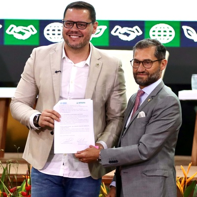 Entrega oficial de documento durante evento institucional, com participantes no palco e painel de identidade visual ao fundo.