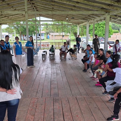 Grupo de pessoas sentadas e em pé em um espaço comunitário coberto, participando de uma atividade coletiva mediada por equipes de apoio, com casas e árvores ao fundo.