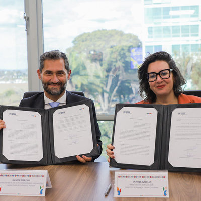 Representante do ACNUR e Ministra dos Direitos Humanos no Brasil estão sentados segurando um acordo assinado nas mãos e sorrido para a foto