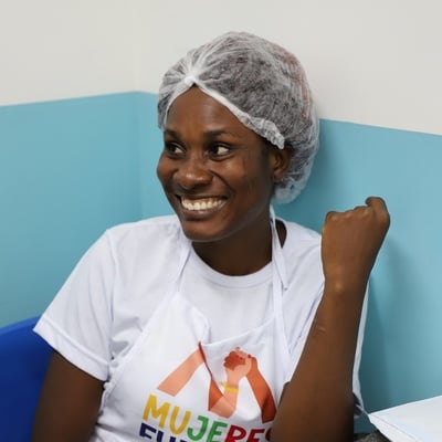 Mulher está sentada frente a uma parede azul e branca. Posa para a foto sorrindo. Ela está vestida com uma camiseta branca com a marca do projeto Mujeres Fuertes. Ela leva touca no cabelo. 
