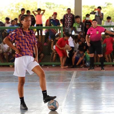 Pessoa participante de uma partida de futsal em uma quadra coberta, com um pé apoiado sobre a bola próximo à linha central. A pessoa veste camisa estampada em laranja e roxo, shorts brancos, meias pretas e chuteiras pretas. Ao fundo, atrás de uma grade verde, há um grande público acompanhando o jogo. Na lateral da quadra, outra pessoa com uniforme rosa está posicionada próxima à área de jogo. A quadra é iluminada pela luz natural, com árvores visíveis ao fundo.