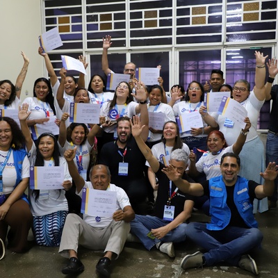 Grupo de participantes do programa Empretec para Refugiados posa com certificados ao final da capacitação, acompanhado por representantes de organizações parceiras, em sala de treinamento.