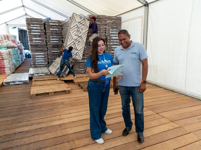 Mulher com uniforme do ACNUR e homem com uniforme da Ortobom verificam papel. Ao fundo, dois homens armazenam colchões em armazém