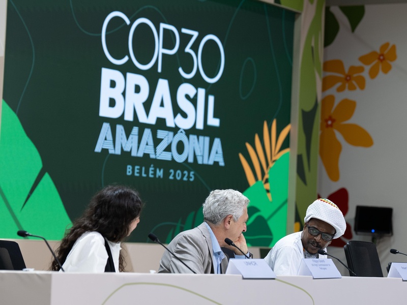 Três pessoas sentadas em mesa de conferência, com painel ao fundo exibindo “COP30 Brasil Amazônia Belém 2025”.