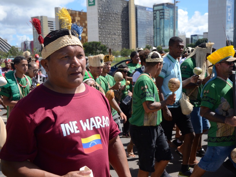 Homem indígena em primeiro plano usa camiseta vinho com o texto ‘INE WARAO’ e uma pequena bandeira estampada, caminhando em meio a um grupo em marcha. Ao redor, várias pessoas vestem camisetas verdes, seguram instrumentos e adornos, com prédios altos ao fundo em uma área urbana.