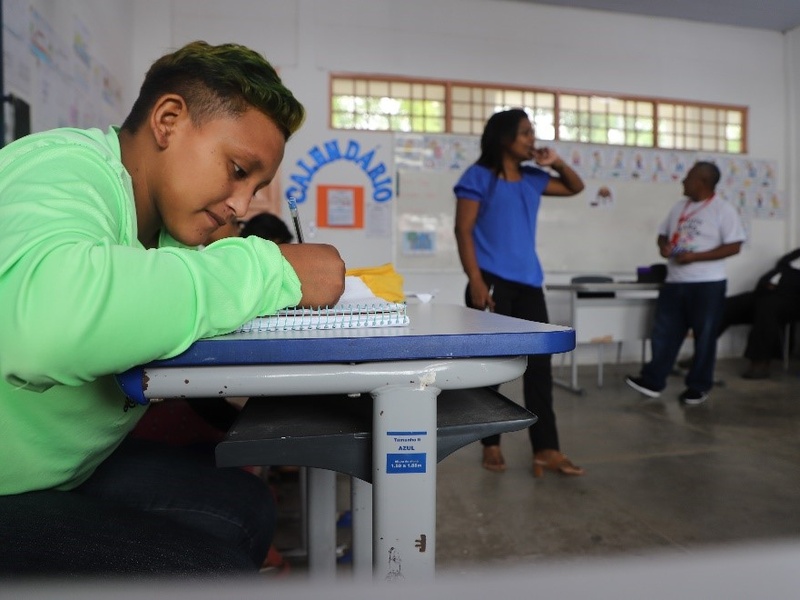 Jovem indígena de blusa verde escreve no caderno numa carteira numa sala de aula. Indígenas Warao frequentam a escola em projeto de alfabetização trilíngue. Em Teresina, crianças e adolescentes de 6 a 17 anos são alfabetizados em português, espanhol e Warao, com o apoio de educadores sociais da etnia indígena.