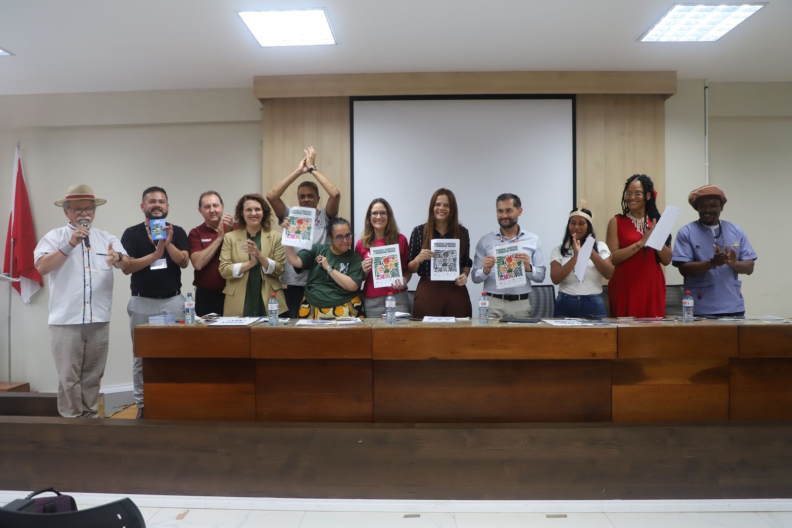 Linha de pessoas em pé segurando cartazes e materiais impressos, em frente a uma mesa de madeira e tela de projeção.