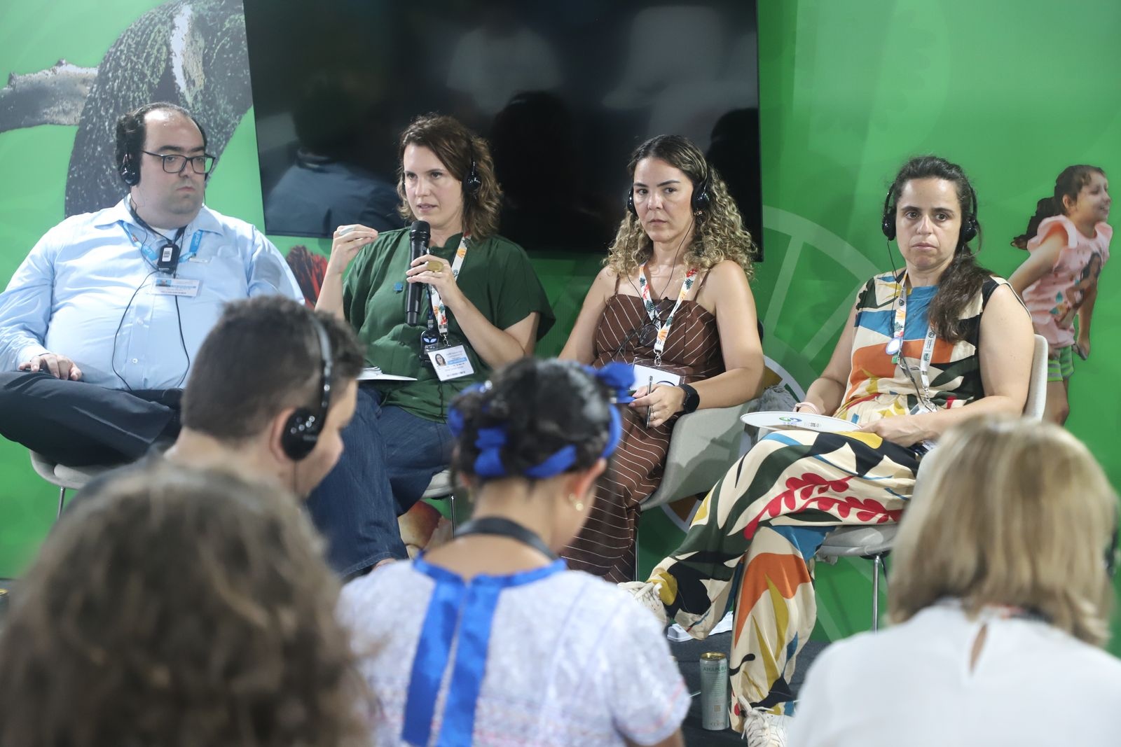 Quatro pessoas sentadas em cadeiras durante um debate, com fundo verde exibindo logotipo das Nações Unidas e ilustrações coloridas..
