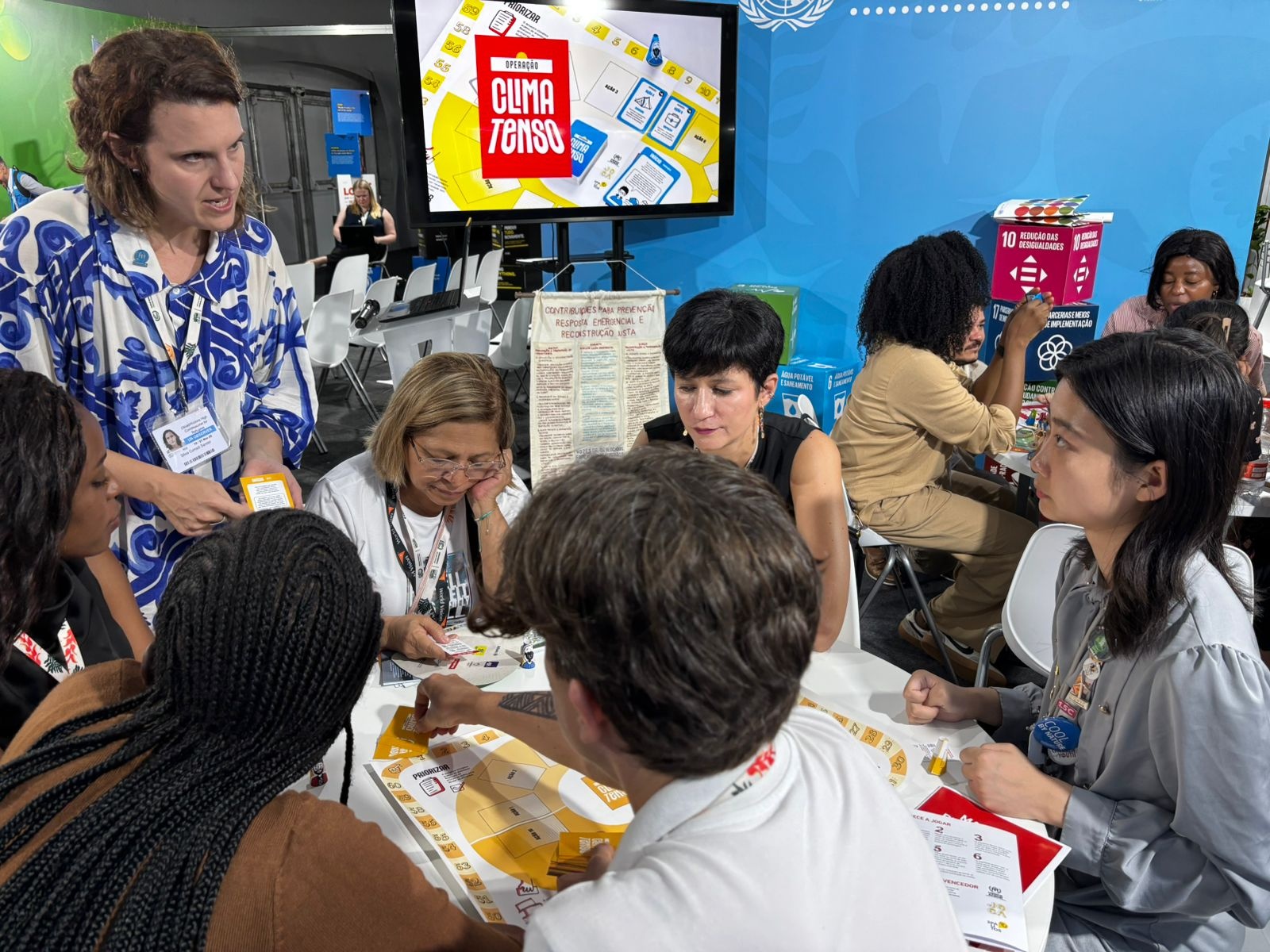 Grupo de pessoas reunidas em torno de uma mesa jogando um jogo educativo, com tela ao fundo exibindo “Clima Tenso”.