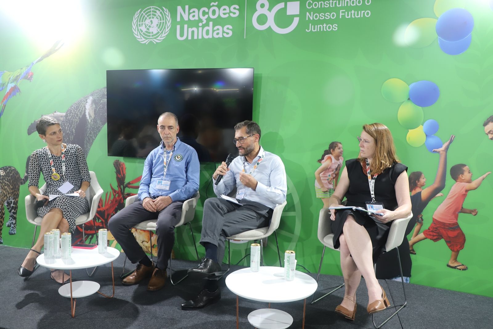 Quatro pessoas sentadas em cadeiras durante um debate, com fundo verde exibindo logotipo das Nações Unidas e ilustrações coloridas.