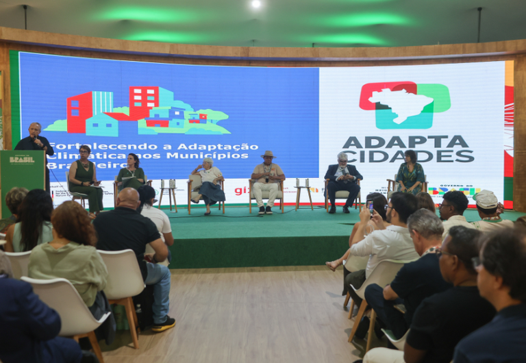 Painel com seis pessoas sentadas em palco, diante de tela grande com logotipo “Adapta Cidades” e texto sobre adaptação climática nos municípios brasileiros.