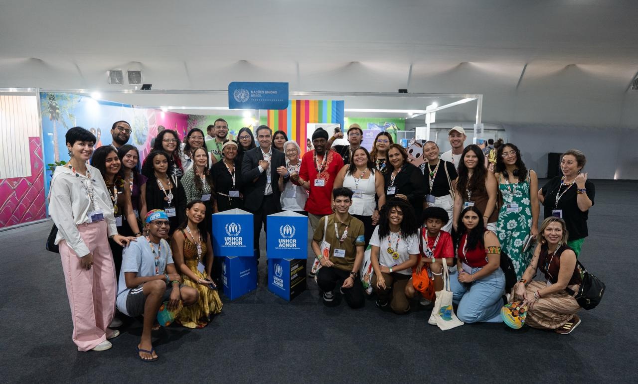 Grupo grande de participantes reunidos em frente a um estande do ACNUR, com banners azuis e decoração colorida ao fundo.