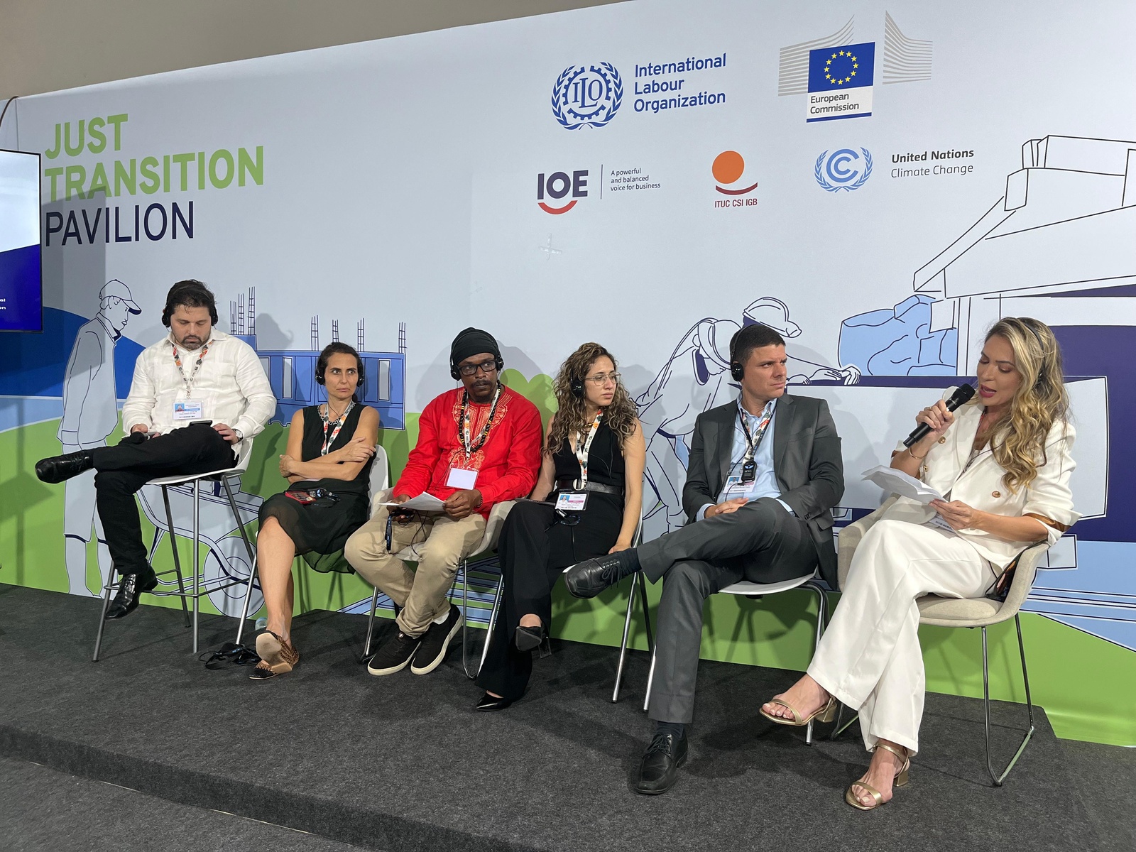Seis pessoas sentadas em palco do Pavilhão de Transição Justa, com logotipos da OIT, Comissão Europeia e ONU ao fundo.