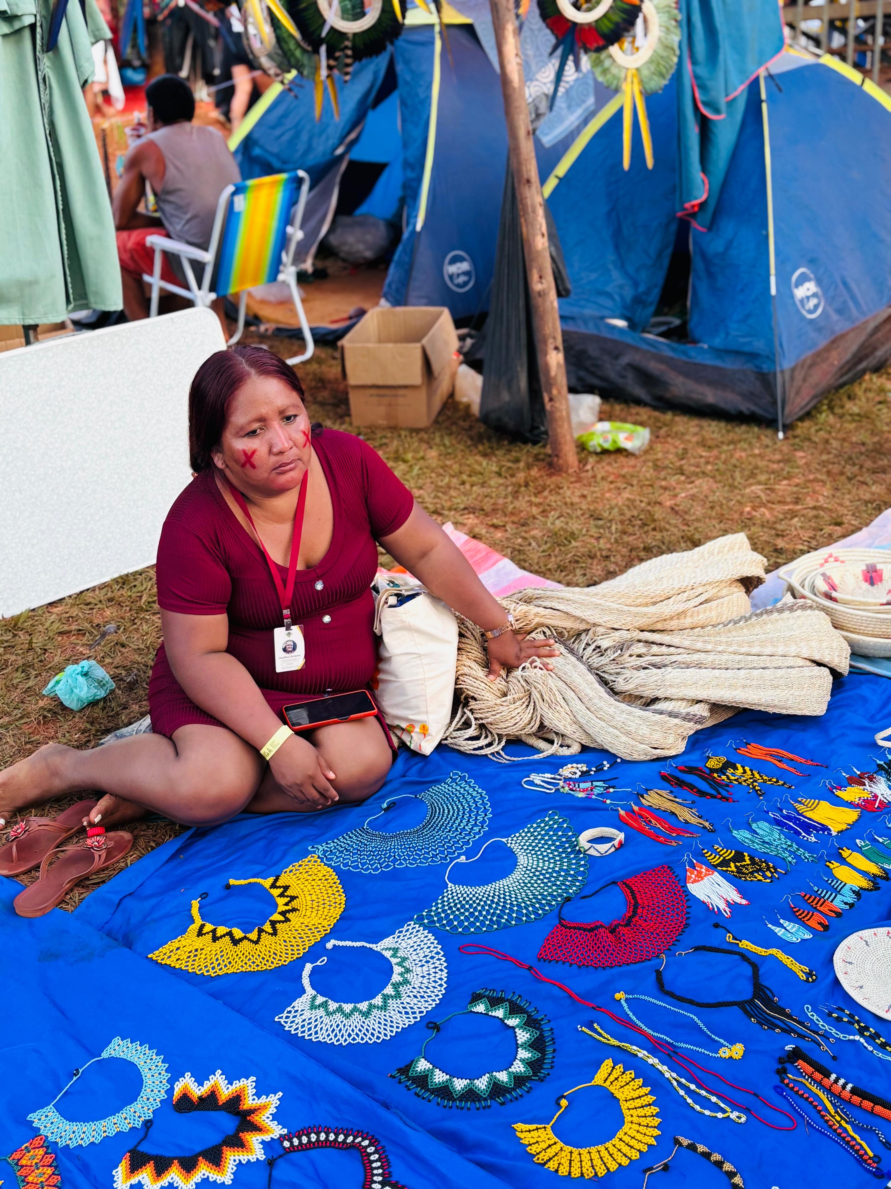 Mulher ind&iacute;gena da etnia Warao est&aacute; sentada em frente a uma barraca de camping, exibindo colares artesanais coloridos dispostos sobre um tecido azul no ch&atilde;o. Ao fundo, h&aacute; outras barracas e pessoas.