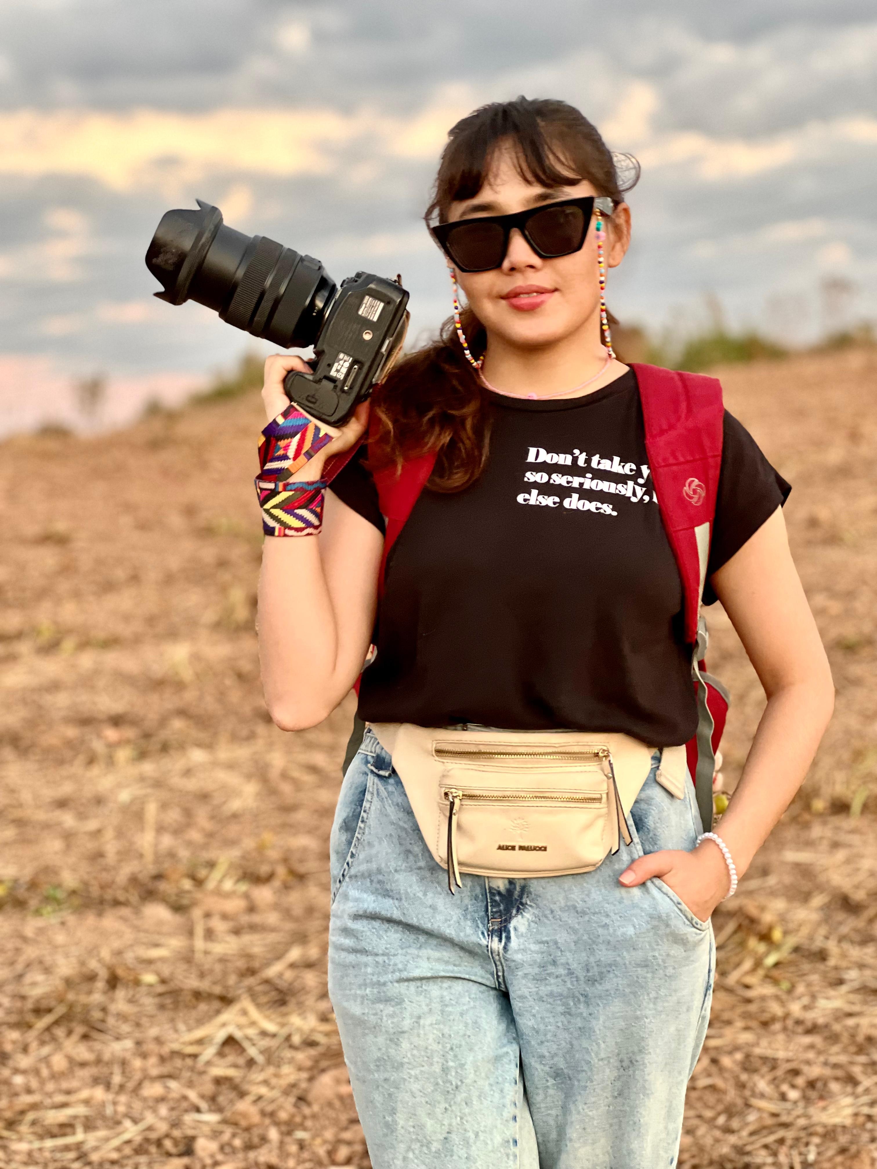 Uma mulher est&aacute; de p&eacute; ao ar livre em um campo, segurando uma c&acirc;mera com uma lente grande na m&atilde;o direita. Ela est&aacute; vestindo uma camiseta preta com texto branco. A pessoa tamb&eacute;m est&aacute; usando jeans azul claro, uma pochete bege na cintura e uma mochila vermelha. O fundo mostra um campo aberto com alguma vegeta&ccedil;&atilde;o e um c&eacute;u nublado.