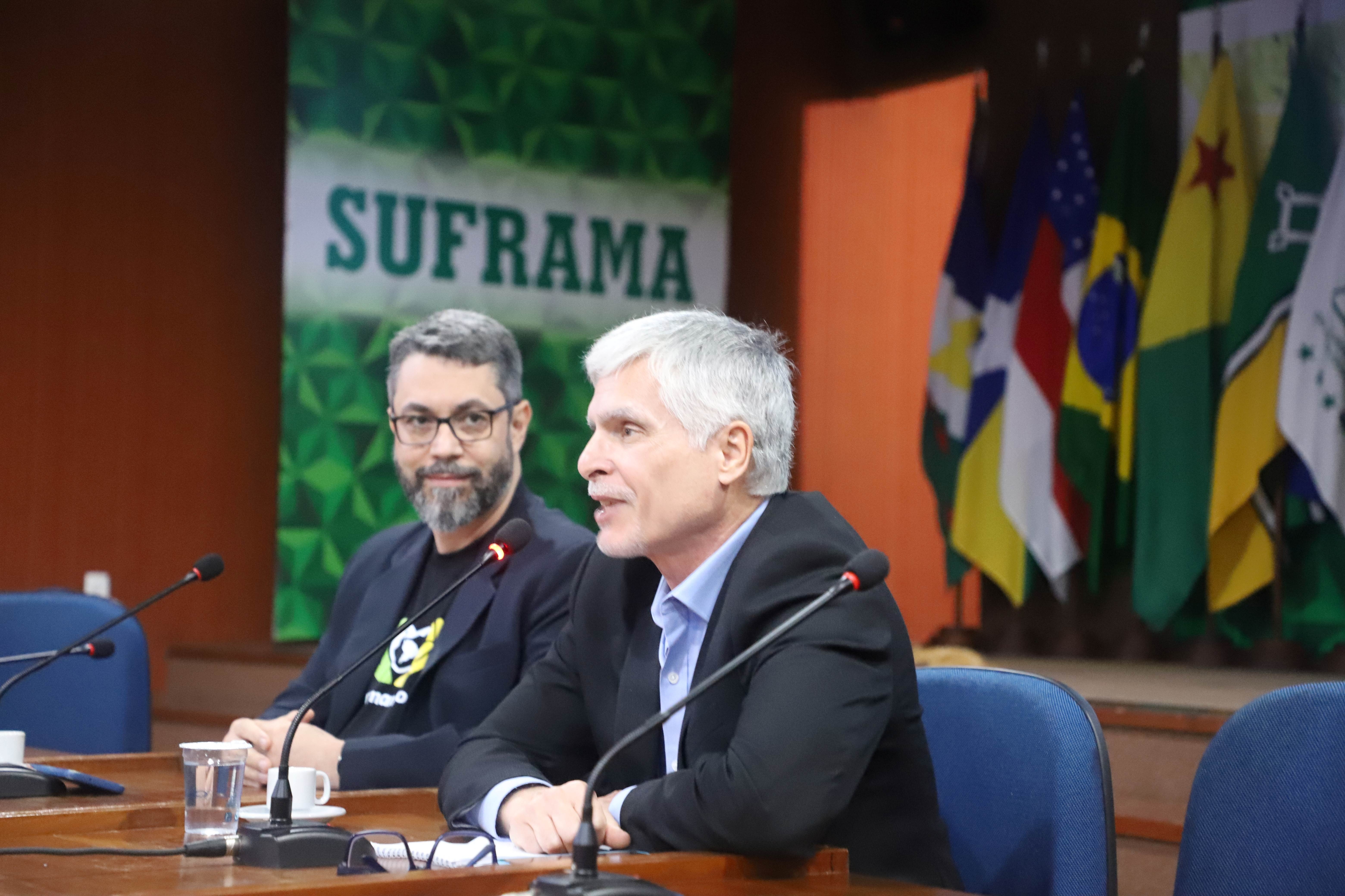 Dois homens brancos sentados em uma mesa. O homem da direita fala ao microfone e atr&aacute;s h&aacute; uma tela escrito Suframa e bandeiras de estados brasileiros. 