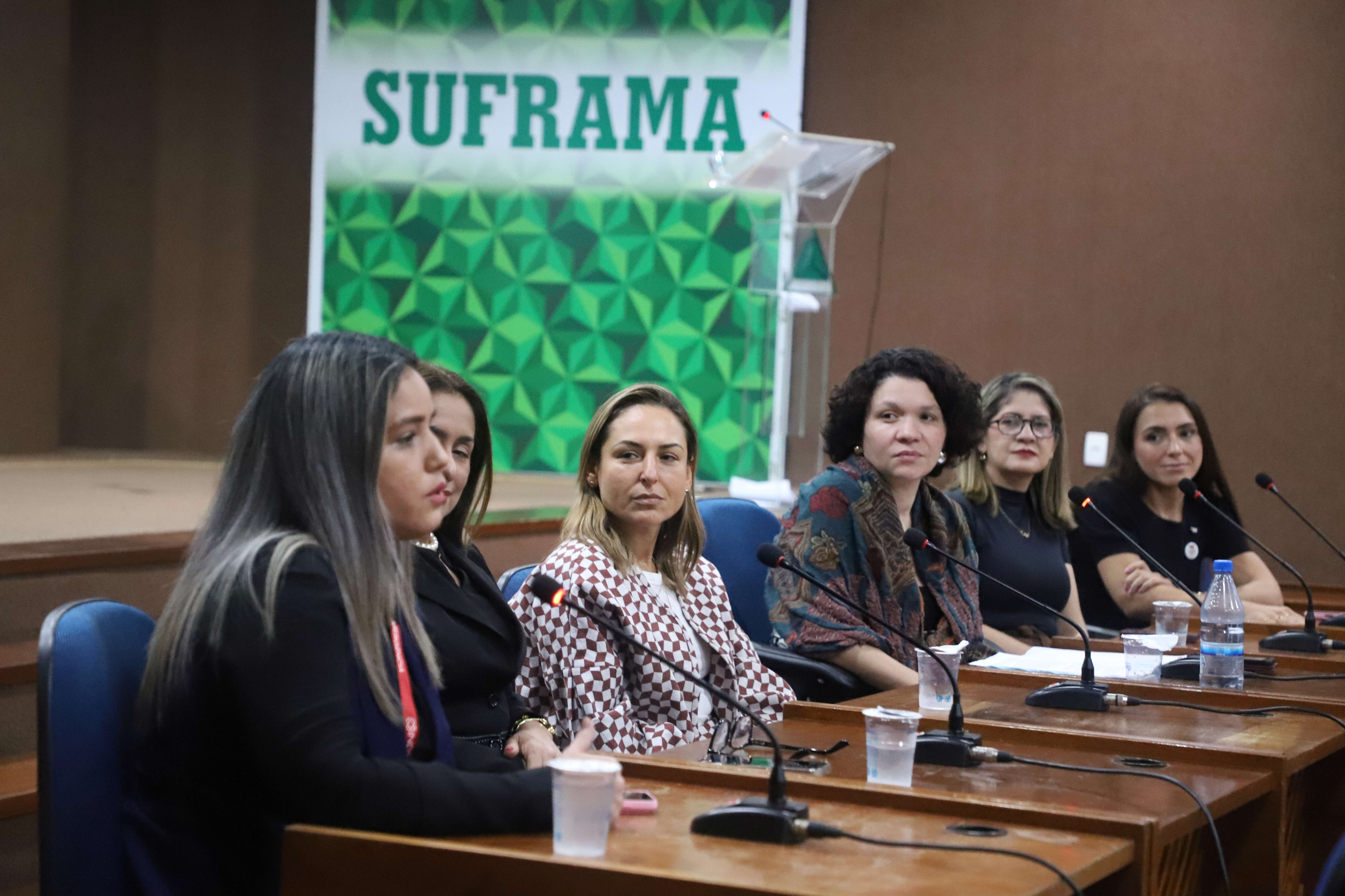 Painel com cinco mulheres sentadas &agrave; mesa com microfones, participando de uma confer&ecirc;ncia. Ao fundo, h&aacute; uma faixa verde e branca com o texto 'SUFRAMA'. Sobre a mesa, h&aacute; garrafas de &aacute;gua e copos.
