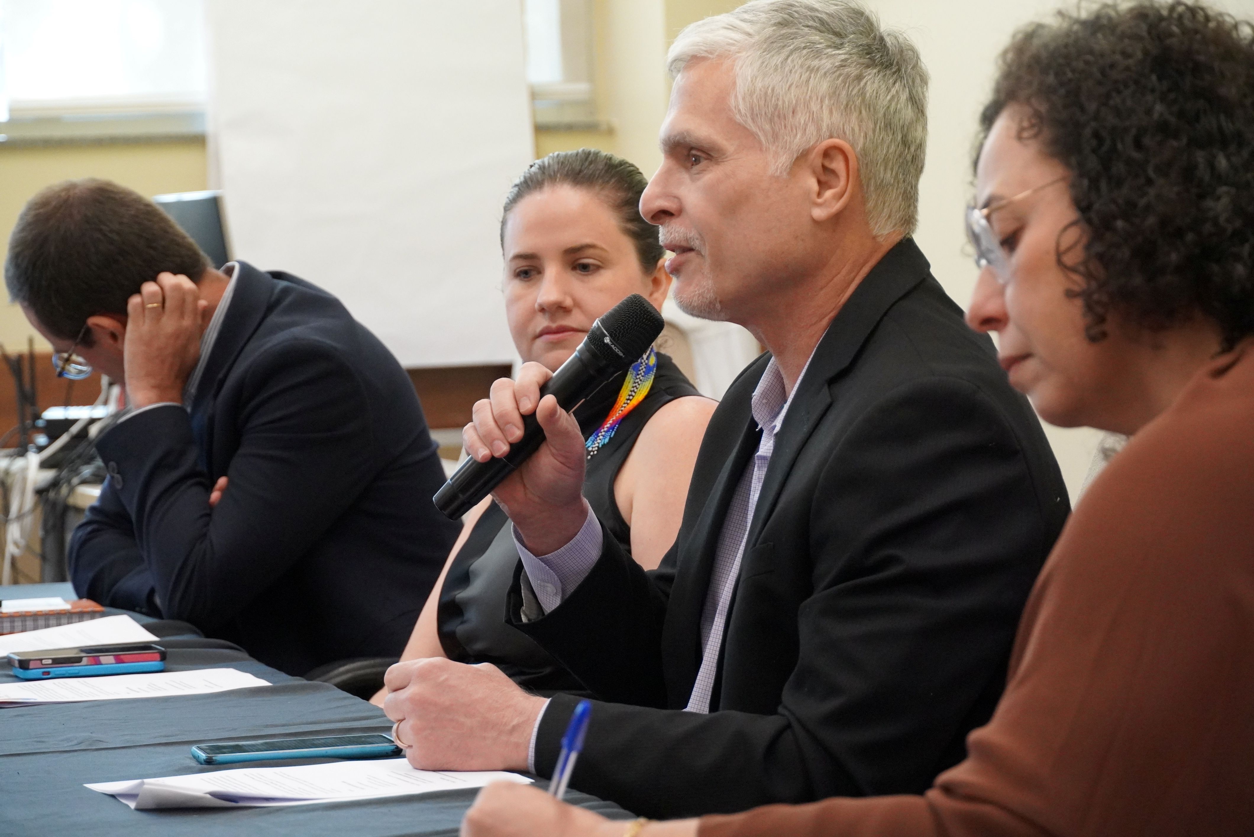 Homem branco sentado entre duas mulheres fala ao microfone