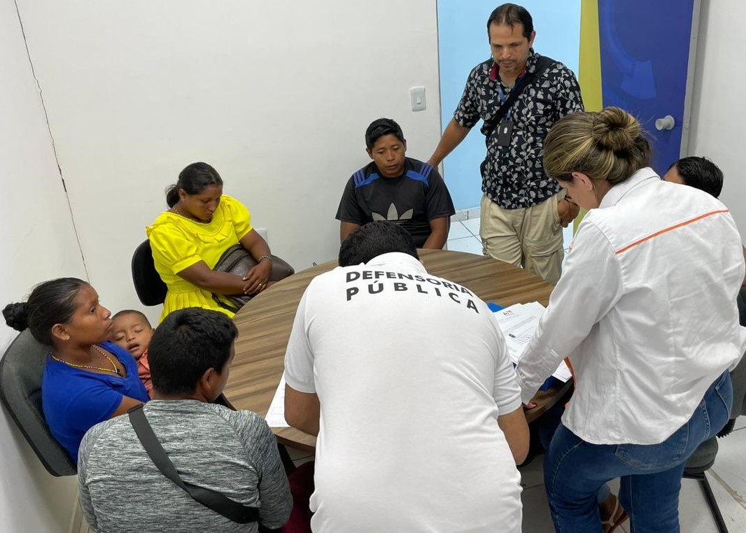 Grupo de pessoas, incluindo ind&iacute;genas da etnia Warao, participam de atendimento com mediador intercultural em uma sala de reuni&otilde;es.