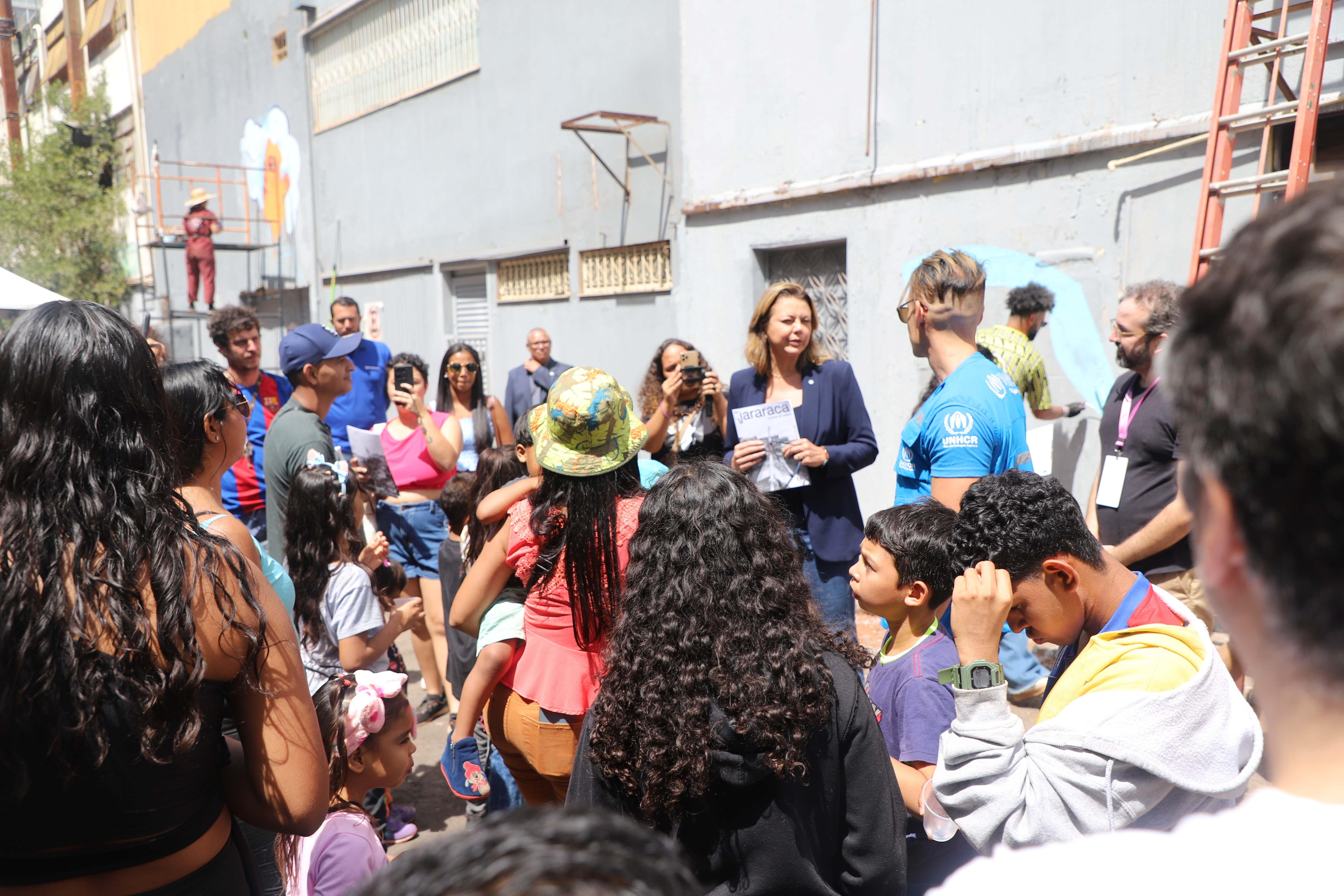 Grupo de adultos e crian&ccedil;as reunidos ao ar livre em frente a um pr&eacute;dio em uma &aacute;rea urbana, com algumas pessoas segurando pap&eacute;is e uma escada vis&iacute;vel ao fundo.
