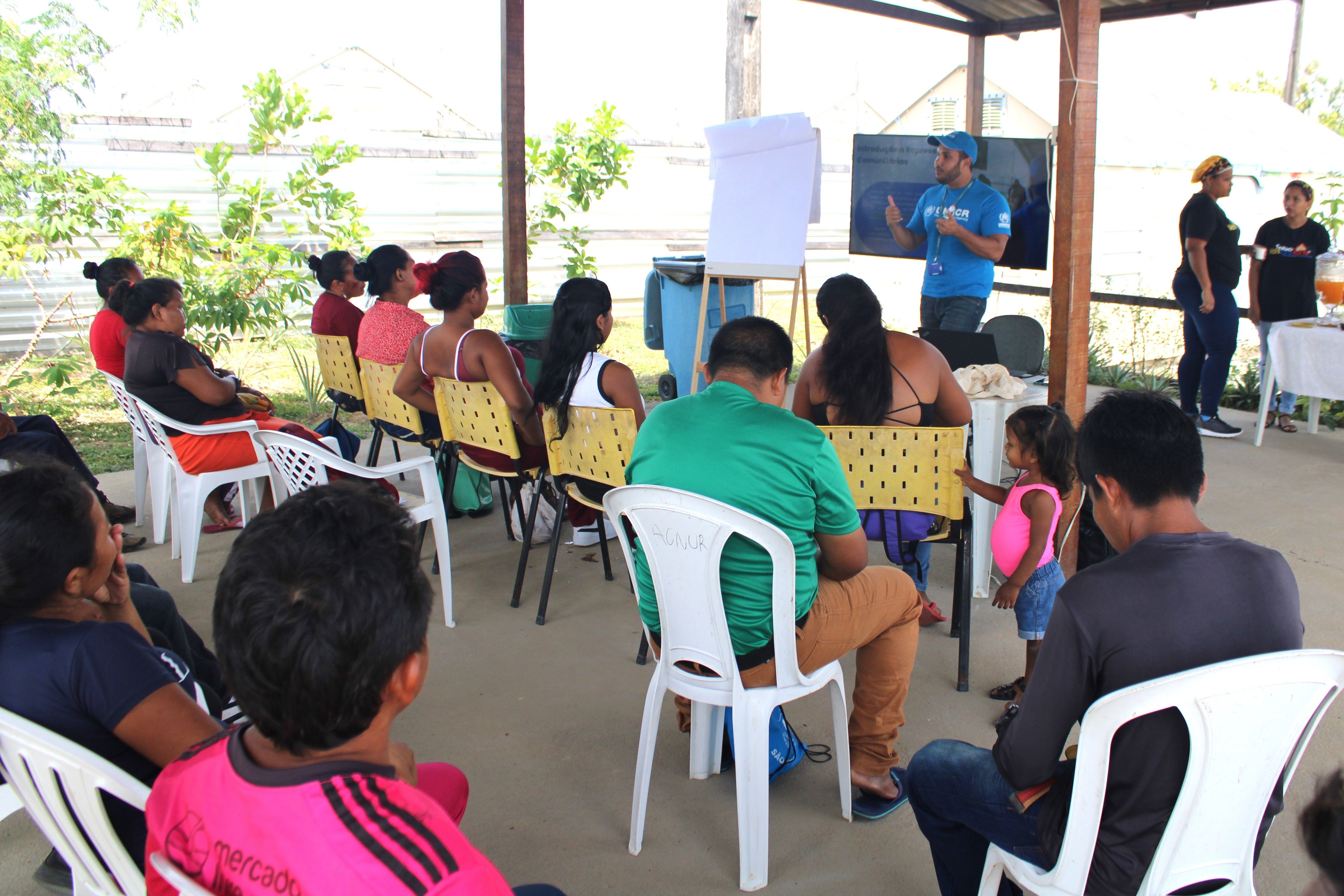 Grupo de pessoas sentadas em ambiente externo, assistindo a uma apresenta&ccedil;&atilde;o. O apresentador est&aacute; em p&eacute; ao lado de uma lousa e uma televis&atilde;o. Algumas pessoas est&atilde;o pr&oacute;ximas a uma mesa com objetos, em um cen&aacute;rio informal e educativo.