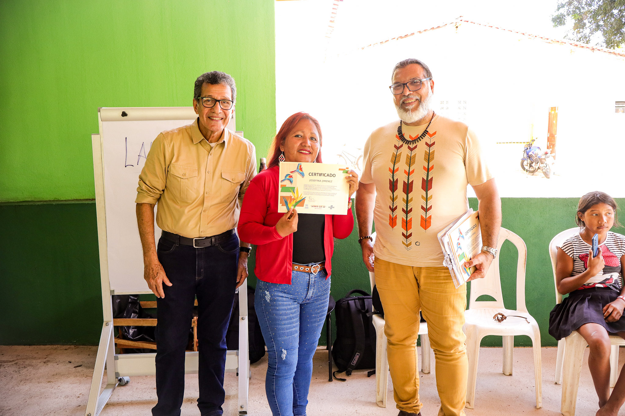 Tr&ecirc;s adultos posam sorrindo para uma foto, com a mulher ao centro segurando um certificado. Eles est&atilde;o em um ambiente interno com paredes verdes e cadeiras brancas de pl&aacute;stico ao fundo.