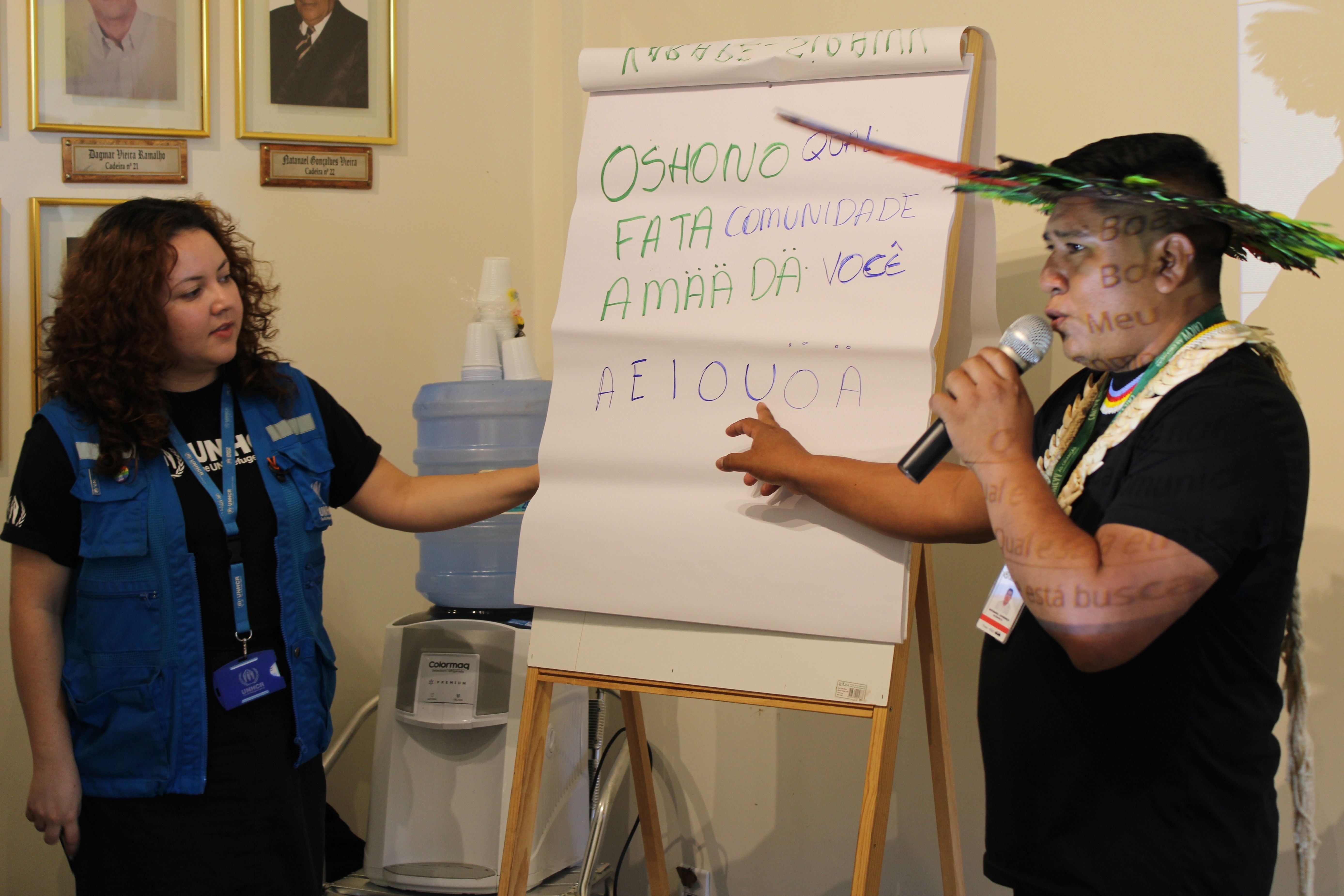 Duas pessoas em p&eacute; ao lado de um flip chart; uma delas segura um microfone e aponta para o quadro, que cont&eacute;m frases escritas em diferentes cores em idiomas ind&iacute;genas.