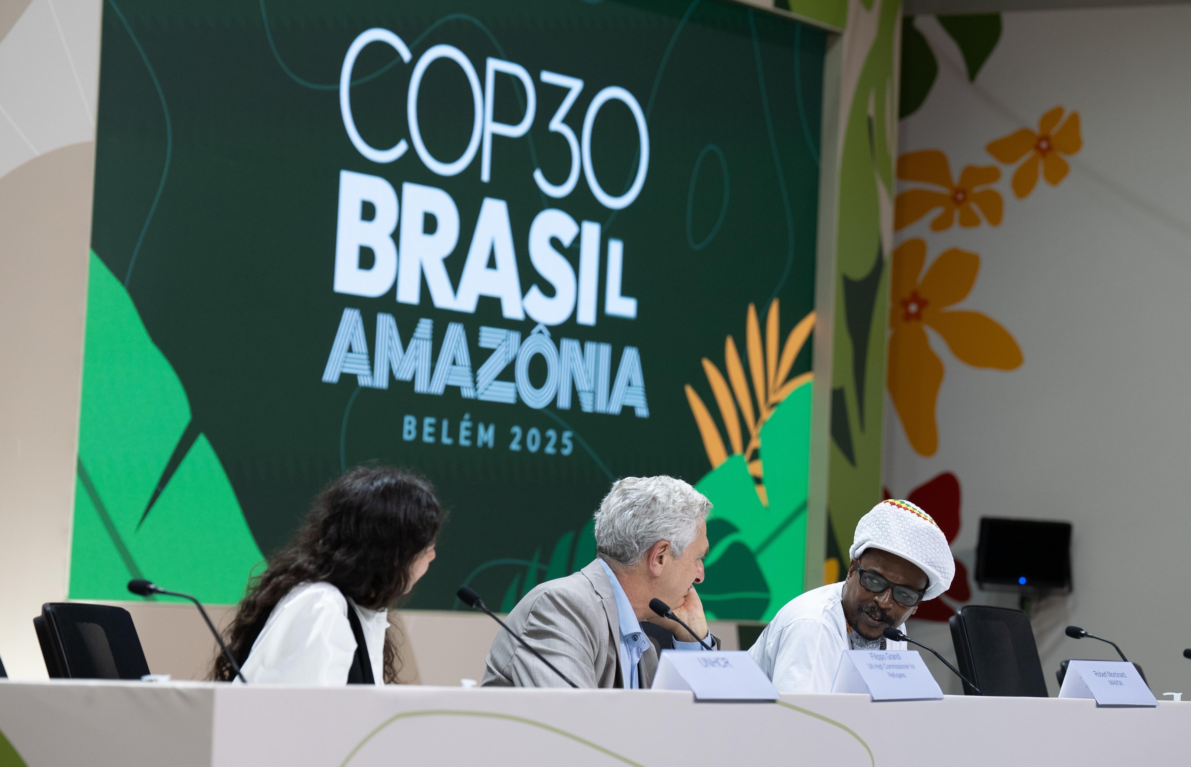 Tr&ecirc;s pessoas sentadas em mesa de confer&ecirc;ncia, com painel ao fundo exibindo &ldquo;COP30 Brasil Amaz&ocirc;nia Bel&eacute;m 2025&rdquo;.