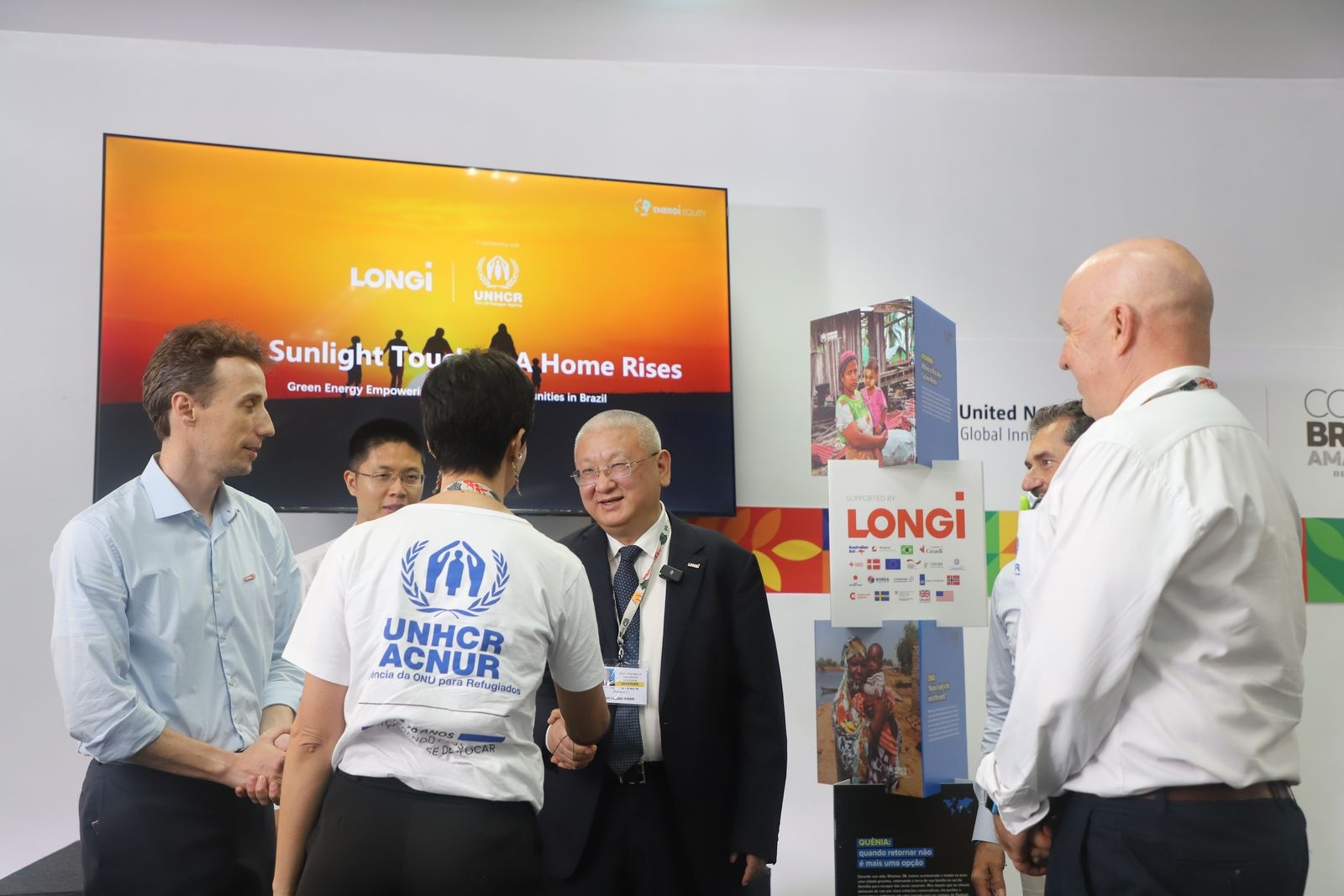 Grupo de pessoas em um estande durante a COP30, diante de uma tela com o t&iacute;tulo &ldquo;Sunlight Touches, A Home Rises &ndash; Green Energy Empowering Vulnerable Communities in Brazil&rdquo;, com os logotipos da LONGi e do ACNUR. Ao lado, h&aacute; um totem expositivo com imagens e informa&ccedil;&otilde;es sobre projetos sustent&aacute;veis. Uma pessoa veste uma camiseta branca com o logotipo do ACNUR e a frase &ldquo;ONU para Refugiados&rdquo;, enquanto cumprimenta outra pessoa. Ambiente interno com identidade visual da confer&ecirc;ncia e materiais promocionais relaci