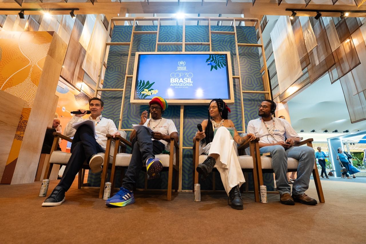 Quatro pessoas sentadas apresentando um painel com uma TV ao fundo com a logo do ACNUR na COP30