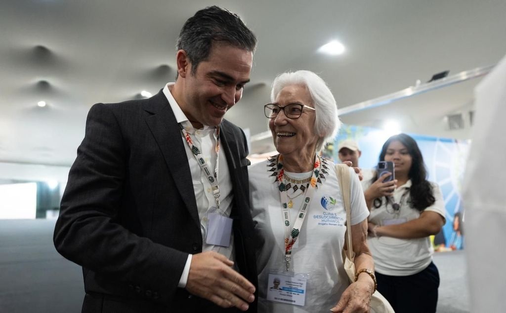 Duas pessoas conversam em um ambiente interno de evento, usando crach&aacute;s de identifica&ccedil;&atilde;o. Uma delas veste terno escuro com camisa clara, enquanto a outra usa camiseta branca com logotipo e colar colorido. Ao fundo, h&aacute; uma pessoa com camiseta branca e um painel azul com elementos gr&aacute;ficos, sugerindo participa&ccedil;&atilde;o na COP30