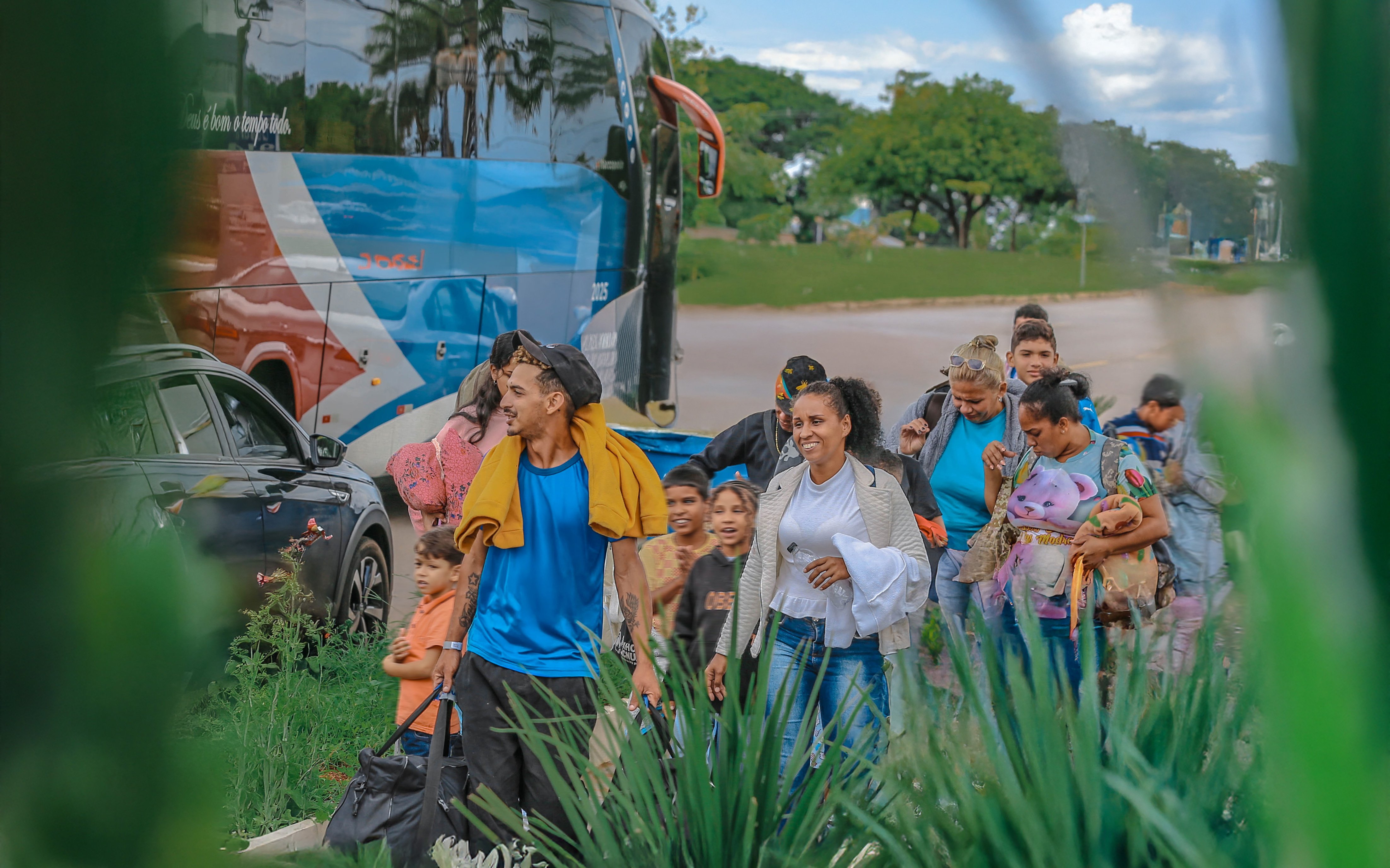 Grupo de pessoas caminhando com malas e crian&ccedil;as pr&oacute;ximo a um &ocirc;nibus estacionado, em uma &aacute;rea aberta com vegeta&ccedil;&atilde;o em primeiro plano e &aacute;rvores ao fundo.