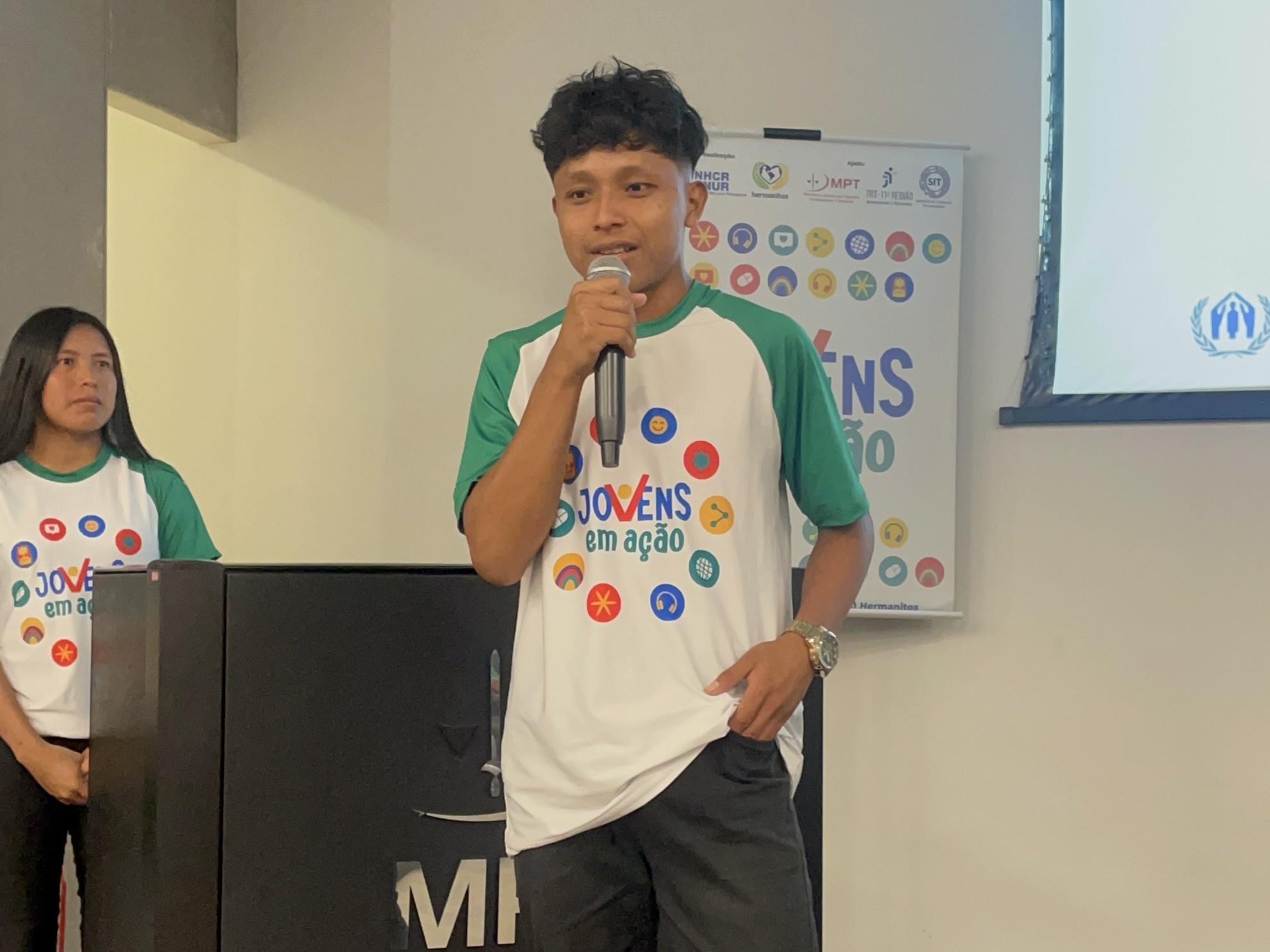 Rapaz jovem segura microfone enquanto fala. Atr&aacute;s dele no canto esquerdo est&aacute; uma jovem que parece assistir o discurso. Os dois vestem camiseta de uniforme do projeto Jovem em A&ccedil;&atilde;o. H&aacute; tamb&eacute;m um banner parcial com a logo do programa. 