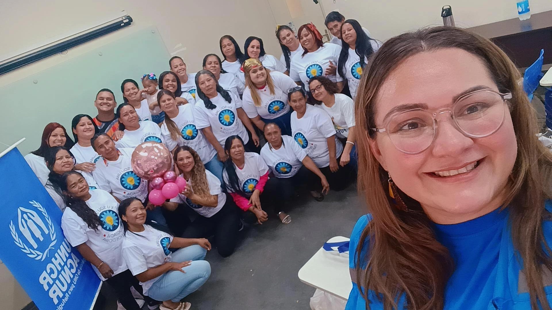 Selfie de uma facilitadora sorrindo &agrave; frente de um grupo de mulheres refugiadas reunidas em uma sala de aula. Todas usam camisetas brancas do curso &lsquo;Eu Sou Uma Mulher Empreendedora&rsquo; e posam juntas, algumas segurando bal&otilde;es rosa. &Agrave; esquerda, h&aacute; um banner azul do ACNUR. O ambiente &eacute; simples, com mesas e quadro branco ao fundo