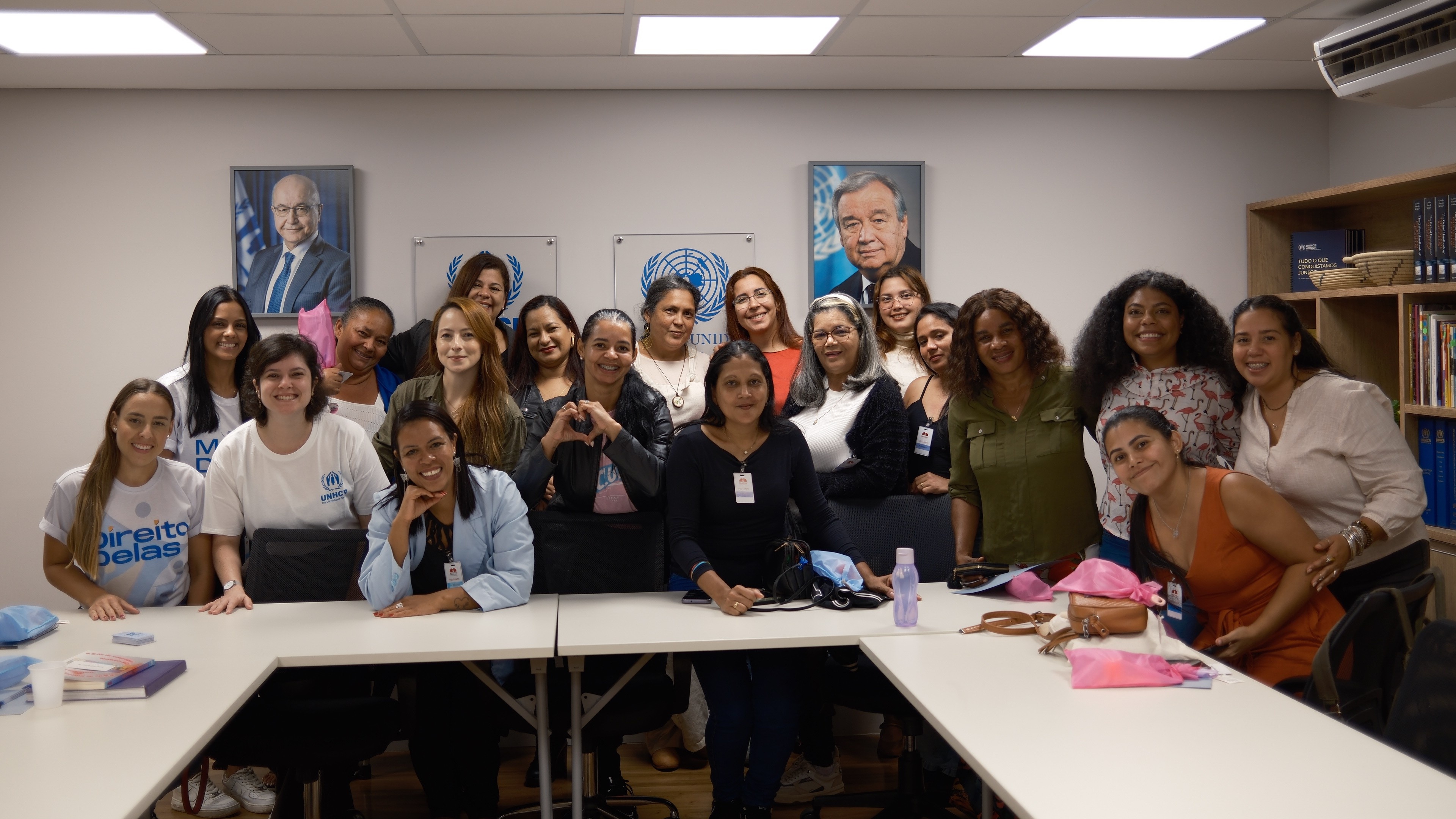 Grupo de mulheres participantes de uma atividade no escrit&oacute;rio do ACNUR em Bras&iacute;lia posa reunido para uma foto em uma sala de reuni&otilde;es. Elas est&atilde;o sorrindo e ocupam o espa&ccedil;o ao redor de mesas brancas. Ao fundo, h&aacute; retratos oficiais e o emblema das Na&ccedil;&otilde;es Unidas na parede, al&eacute;m de estantes com livros. Algumas seguram lembran&ccedil;as e materiais distribu&iacute;dos durante o encontro