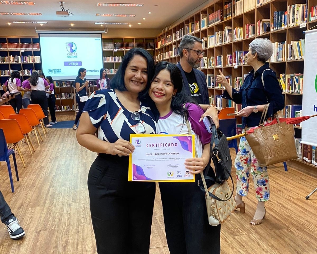 Duas mulheres seguram um certificado. Ao fundo, uma biblioteca comp&otilde;e o cen&aacute;rio. 