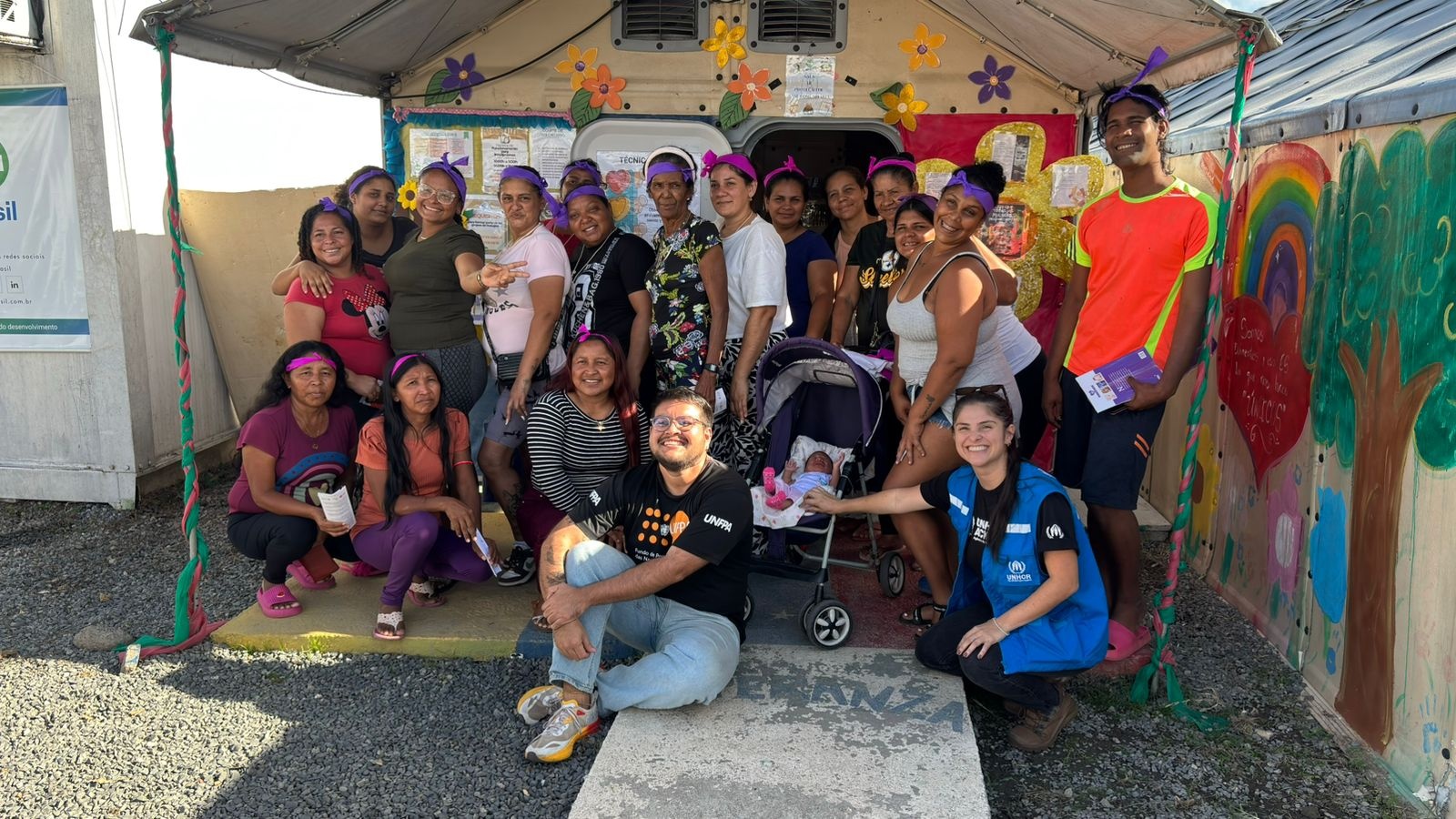 Grupo de mulheres refugiadas e membros de organiza&ccedil;&otilde;es parceiras posam em frente a uma estrutura decorada com flores e pinturas coloridas durante uma atividade do Dia Internacional da Mulher. As participantes usam viseiras roxas e sorriem para a foto; ao centro, h&aacute; um carrinho de beb&ecirc;. A cena ocorre em &aacute;rea externa, com murais pintados &agrave; m&atilde;o ao fundo e clima de celebra&ccedil;&atilde;o comunit&aacute;ria.